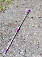 Long metal handled cleaning tool with purple plastic components, laid on gravel surface