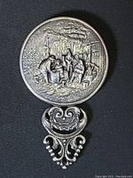 Front side of the round pocket mirror showing a detailed raised scene of people gathered outdoors with fine detail.