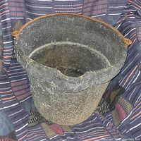 Black heavy duty rubber farm bucket with textured, worn surface showing dirt and hay remnants. Thick rope handle tied through holes near top edges visible.