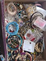 Box filled with assorted costume jewelry items including bracelets, necklaces, rings, and earrings in various colors and materials like beads, faux pearls, and metal.