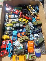 Top-down view of a large cardboard box filled with approximately 40 assorted metal die-cast toy vehicles including cars, tanks, trucks, and boats in multiple colors and worn condition.