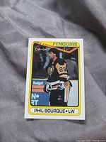 Front view of Phil Bourque hockey card showing player in Penguins uniform, O-Pee-Chee branding, yellow border, and player name/position details.