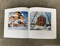 Open book showing full color paintings of snow-covered houses, showcasing the vibrant and detailed artwork of the Group of Seven.