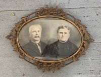 Photo showing large oval ornate brass frame with convex glass featuring a black-and-white vintage portrait of a man and woman.