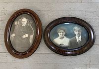 Pair of large antique oval wooden frames with convex glass, each containing black and white vintage portraits. The left frame shows a single man seated in a chair, the right frame shows an elderly couple. Both frames are on a light wooden floor.