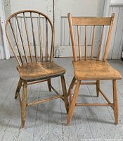 Two antique wooden side chairs placed side by side on a wooden floor showing differing backrest designs and wood finish.