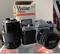 Asahi Pentax K1000 camera with SMC Pentax 55mm prime lens mounted, 80-200mm zoom lens beside it, and Vivitar 2800 flash with original box behind.