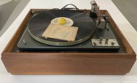 Front view of turntable showing wooden plinth, platter with adapter and manual envelope, tonearm assembly