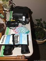 Photo showing Tasco binoculars, Olympus and Minolta film cameras, black carrying case and camera accessories arranged on white table