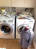 Wide shot showing stack of plastic and fabric hangers on top of front-loading washer and dryer, dress form mannequin posed with striped shirt in front.