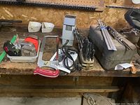 Photo showing measuring tapes, Black and Decker drill bit sharpener, hacksaw, some drill bits, and the metal toolbox on a wooden workbench.