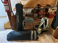 Lot of fishing rods in wall frame, green tent bag, folding chair, oar, folding stools, and lantern arranged on floor near basement stairs