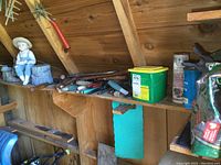 Shelf displaying assorted gardening tools and fuel cans along with a figurine of a boy with a straw hat