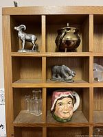 Wooden display case with multiple compartments holding various miniatures including a Toby jug, brass figurines, glass bottles, and animal figures