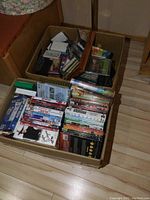 Top view of a cardboard box filled with DVDs, VHS tapes, CDs, and cassettes showing a variety of items in mixed genres.
