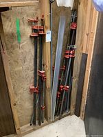 Several pipe clamps of varying lengths, metal pipes with red clamp heads, organized vertically in a corner with some wall boards behind
