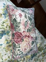 Close-up of a rectangular cushion with floral fabric featuring pink, cream, and green roses and leaves, with pink piping around edges, placed on a floral-patterned surface.