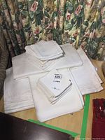 Stack of folded white patterned table linens including tablecloths and napkins on a wooden surface with floral curtain background.