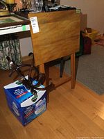 Wood card table folded, showing top surface and legs; wooden coat rack seen partially; Vicks humidifier box on floor