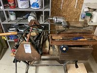 Overall shot showing wooden tool boxes with various tools including manual drill and files, piled on a metal cart in a basement workspace.