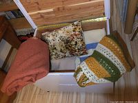 Open white wooden blanket box containing folded blankets, crocheted afghan throw draped over side, and floral cushion inside.