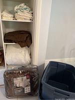 Photo of the shelving unit holding folded white and brown textiles, pillow cases and shams stacked on the upper shelves, and a plastic bin with an 8 piece brown complete bed set package on the floor.