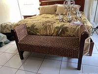 Full front and side view of the woven seagrass and wood bench placed near a bed, showing the seat, backrest, and wooden legs.