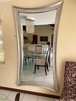 Full view of a large rectangular mirror with a curved silver wood frame mounted on a wall reflecting a room with table and chairs.