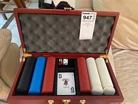 Open wooden poker case showing four stacks of poker chips in black, blue, red, and white, two dice, and a deck of playing cards with Joker card on top.