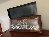 Photo of the framed zebra print on a wall below a mounted TV, showing the full item and wooden frame with textured and patterned design.
