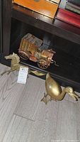 Photo of brass duck, brass horse head sculpture, and vintage brass and copper car music box on wooden floor and shelf.