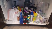 Wide view of the cabinet under the sink showing various cleaning supplies and plastic kitchen waste bags stored inside.