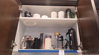 Full view of cabinet shelf with coffee and tea items including mugs, coffee maker, grinder, French press