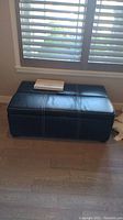 Black faux leather rectangular ottoman with white stitching, placed against window.