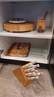 Overview on shelf and floor showing knife block with knives, wood and glass cutting boards on shelf, round chopping block and mortar & pestle above.