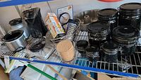 Wide view of black canisters with lids, metal bundt pan, grater, cork coasters, and various kitchen items on metal shelving.