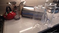 Photo of red whistling kettle, acrylic pitchers, metal breadbox, and utensil pail with kitchen tools on countertop.