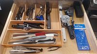 Full view of kitchen drawer with knives, forks, pizza cutters, tongs, and treat bags.