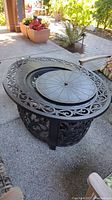 Front-left perspective of oval cast aluminum table showing central burner with circular lid and decorative scrollwork.