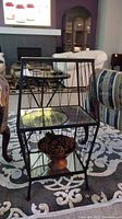 Side view showing black metal plant stand with glass top and mirrored shelves, wooden bowl with pinecones on bottom shelf.