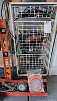 Full view of three white wire baskets filled with assorted hand and power tools and accessories on a cart, plus a ladder and a Black & Decker grinding wheel pack.