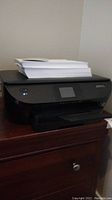 Black HP Envy 5600 printer with touchscreen and stack of white paper on top.