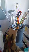 Photo capture of yard care tools including crutches, brooms, snow brushes, and squeegee inside a gray Brute container in a storage or garage area.