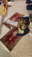 Four area rugs displayed on carpeted floor, two unrolled matching red floral rugs, one rolled blue floral rug, and one decorative tapestry.