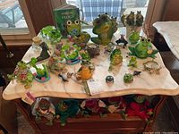 Photo showing a marble table top with about 20 frog collectibles including ceramic frogs, glass frog figurines, small decorative items, and a frog-themed book.