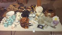 Full view of table with assorted glassware, china, mugs, wood trays, cake container and other kitchen and decorative items arranged.