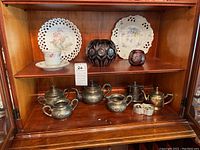 Full view of bottom two shelves of cabinet showing assortment of porcelain plates on upper shelf, cranberry cut glass vases, and silver plate tea set arranged on lower shelves.