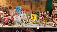Overview of assorted vintage Christmas decorations on tabletop including plush elves, ceramic vases, blow mold Santa head, signs and ornaments