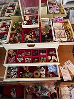 Full view of cream-colored jewelry box with three red-velvet lined drawers opened, containing assorted brooches, necklaces and earrings.