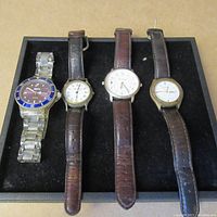 Four vintage watches displayed in a row on a black tray. Three have brown leather bands, one has a silver metal band. Watches show signs of wear.
